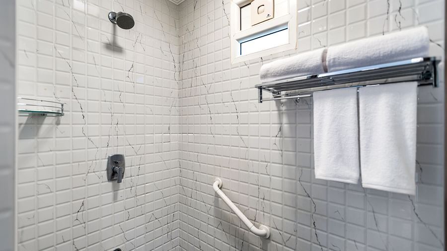 A shower stall with white square tiles, a shower head, a grab bar, and a towel rack with folded white towels - Mango Hill Resort, Coimbatore