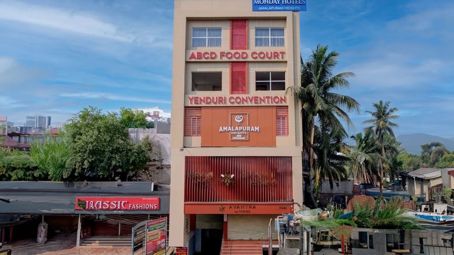 Exterior front view of Monday Hotels Amalapuram Heights building surrounded by trees under a clear blue sky.