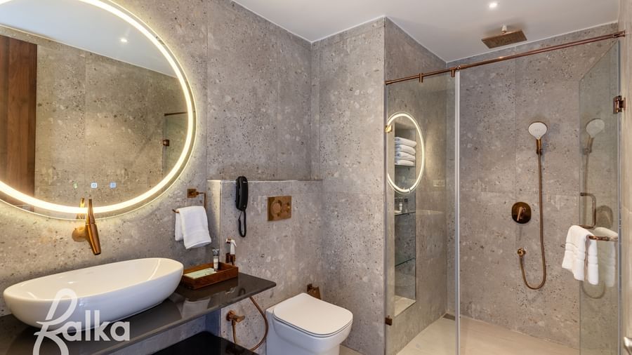 A modern bathroom at Osian Valley Resort & Spa with a stone basin on a dark counter, a large illuminated mirror, and a walk-in shower with a glass screen.