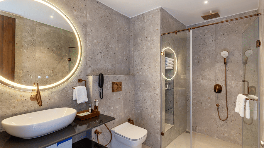 A modern ensuite bathroom at Osian Valley Resort & Spa, Kumbhalgarh, equipped with a walk-in shower, round backlit mirror, stylish wash basin, and neatly arranged bath amenities.