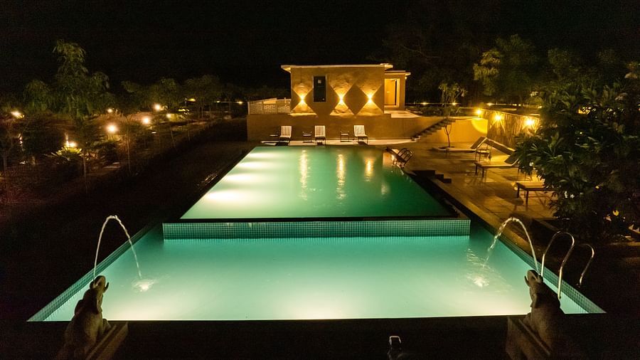 A serene night time view of a shimmering swimming pool illuminated by soft lighting, surrounded by lush trees - Rajbagh Wellness, Udaipur