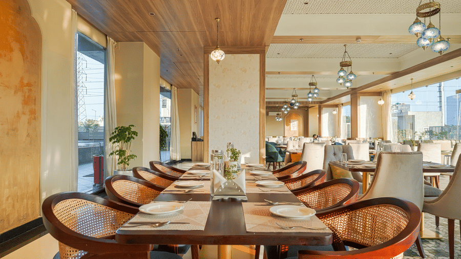 A dining space at Urban Rasoi at Urban Suites by BluSalzz, Jaipur, with wooden tables and chairs, set for a meal, with ample natural light from large windows.