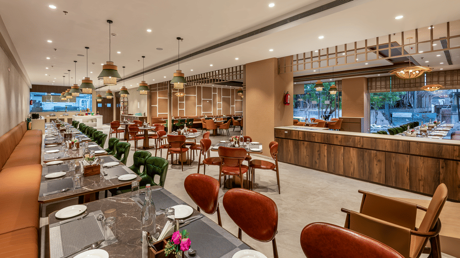  The spacious and modern interiors of the Blue Coriander restaurant featuring emerald green and mustard yellow chairs, marble-topped tables, and large woven pendant lights – Skkylight Lords Inn, Nanded