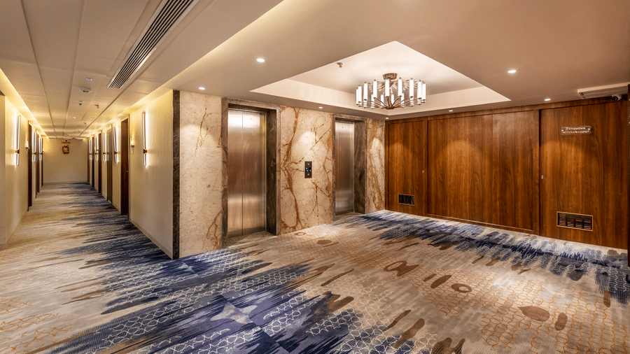 A modern lift lobby with two stainless steel elevators set in marble walls, wood panelling, a small chandelier, and a patterned blue carpet - Skkylight Lords Inn, Nanded