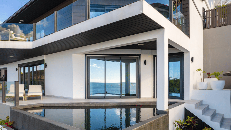  A front view of a two story villa at Tamarind Hills Resort and Villas, Antigua, featuring two lounge chairs, expansive glass windows, and a private infinity-edge pool.