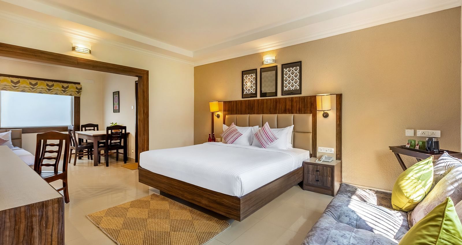 A hotel room with a queen bed, a sofa, and a doorway leading to a dining area with a table and chairs - Mango Hill Resort, Coimbatore