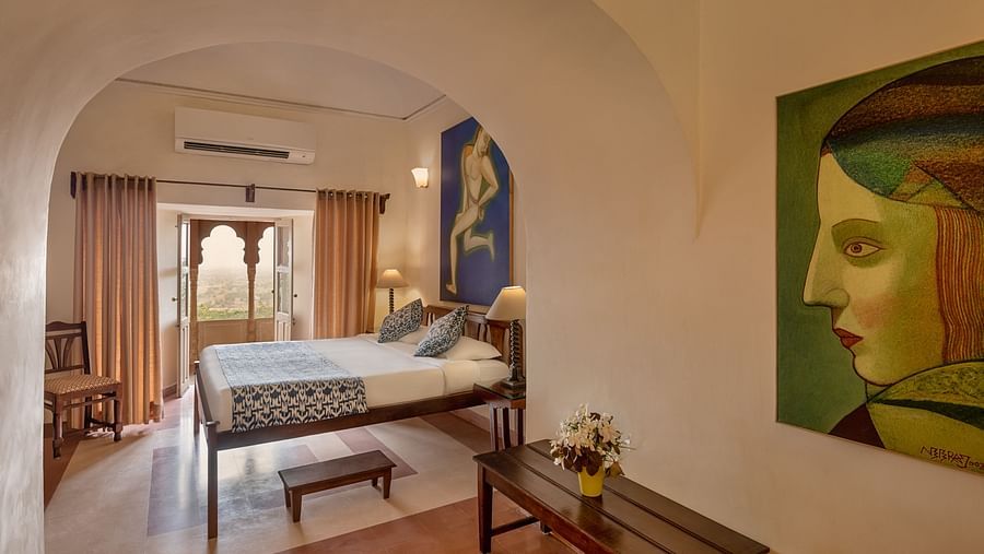 A room with a bed, a bench, a framed artwork, and an arched doorway leading to an outside view in Tijara Fort-Palace - 19th Century, Alwar.