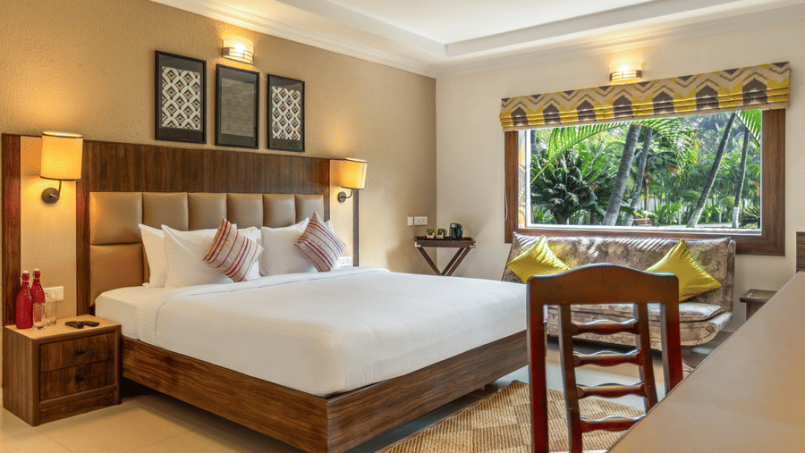 A hotel room with a bed, a desk, a sofa, and a large window overlooking a garden with palm tree at Mango Hill Resort, Coimbatore