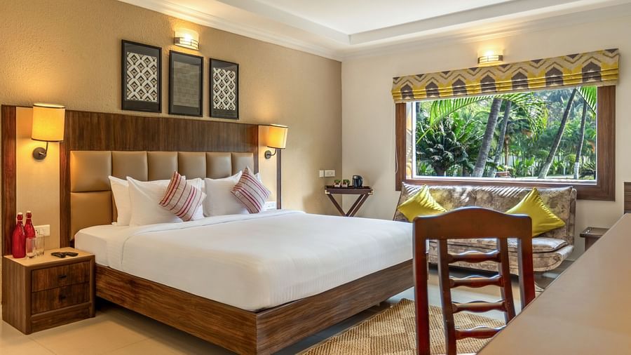 A hotel room with a bed, a desk, a sofa, and a large window overlooking a garden with palm tree - Mango Hill Resort, Coimbatore