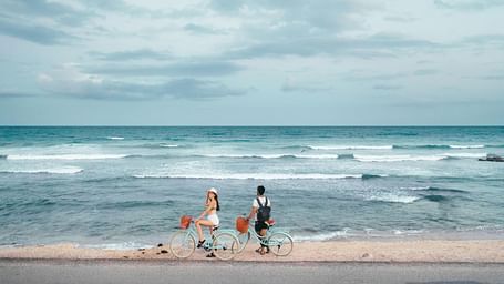 A wide-angle view of the beach with two people standing beside their bicycles, gazing at the calm sea under a clear sky.