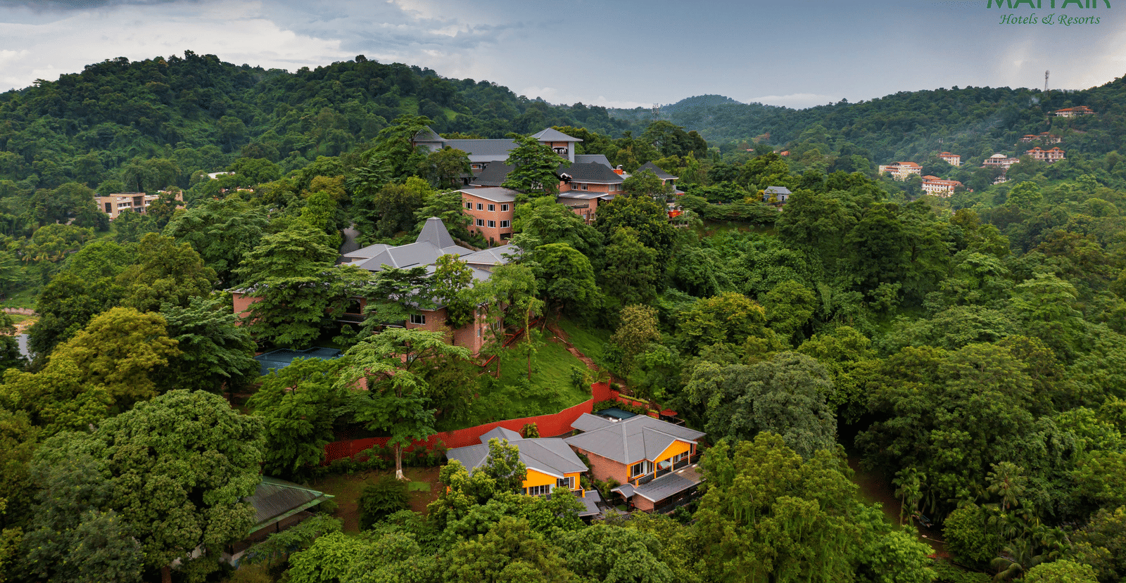 Aerial view of MAYFAIR Spring Valley Resort Guwahati surrounded by trees with a backdrop of mountains under a cloudy sky