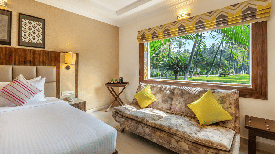 A hotel room with a queen bed, a patterned sofa, and a large window overlooking a garden with palm trees - Mango Hill Resort, Coimbatore