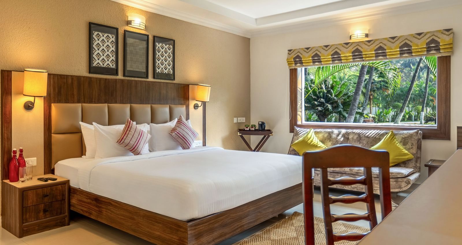 A hotel room with a bed, a desk, a sofa, and a large window overlooking a garden with palm tree - Mango Hill Resort, Coimbatore