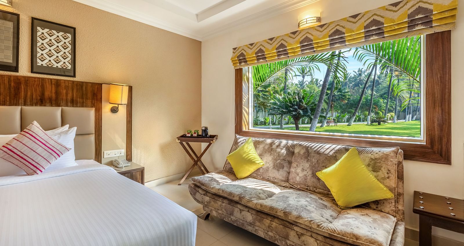 A hotel room with a queen bed, a patterned sofa, and a large window overlooking a garden with palm trees - Mango Hill Resort, Coimbatore