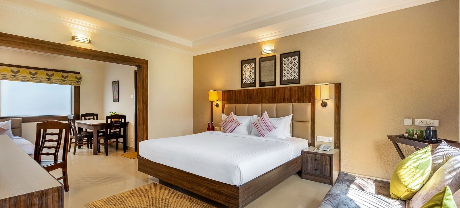 A hotel room with a queen bed, a sofa, and a doorway leading to a dining area with a table and chairs - Mango Hill Resort, Coimbatore