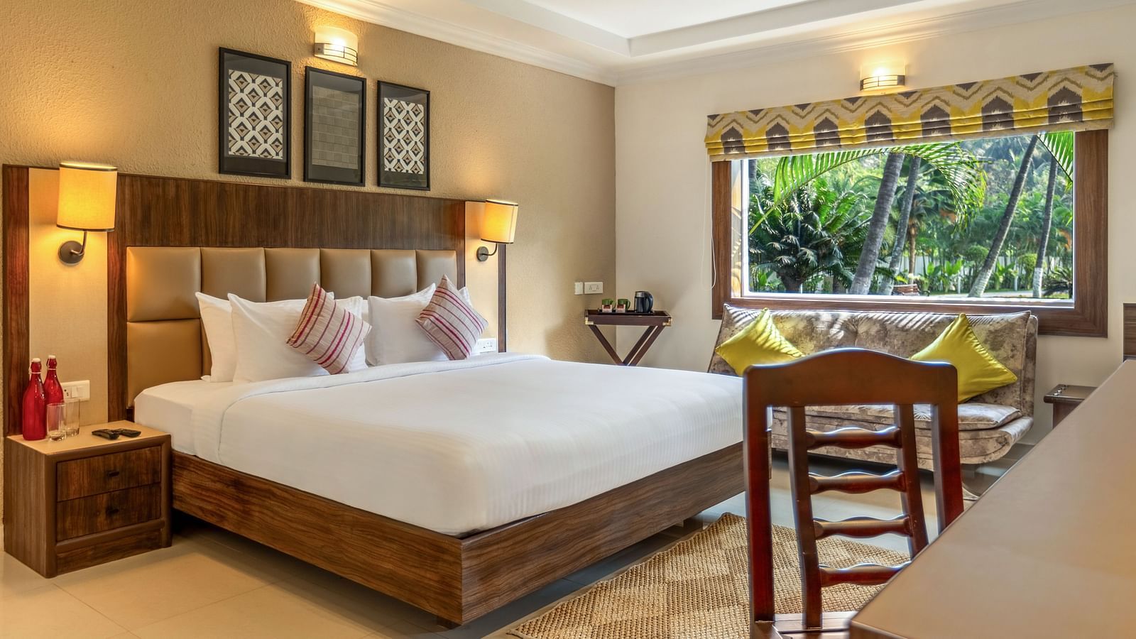 A hotel room with a bed, a desk, a sofa, and a large window overlooking a garden with palm tree - Mango Hill Resort, Coimbatore