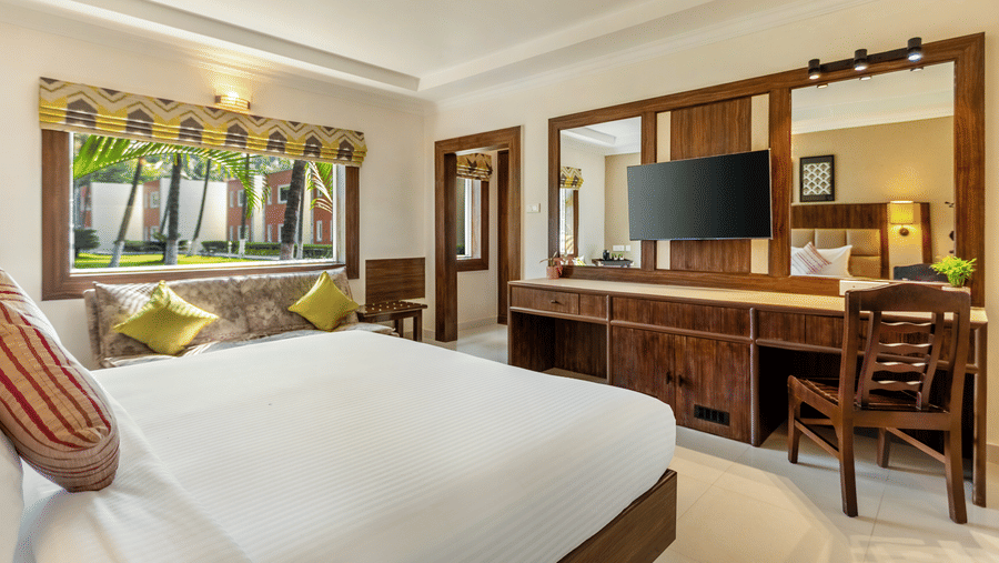 A bright bedroom with a bed with white linens, a wooden desk with a mounted TV, a patterned sofa with yellow pillows, and a window view of a tropical garden - Mango Hill Resort, Coimbatore