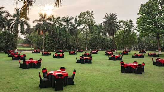 Outdoor lawn setup with round tables covered in red and black linen, surrounded by palm trees and greenery under a cloudy sky at DGV Resort by Treat, Silvassa