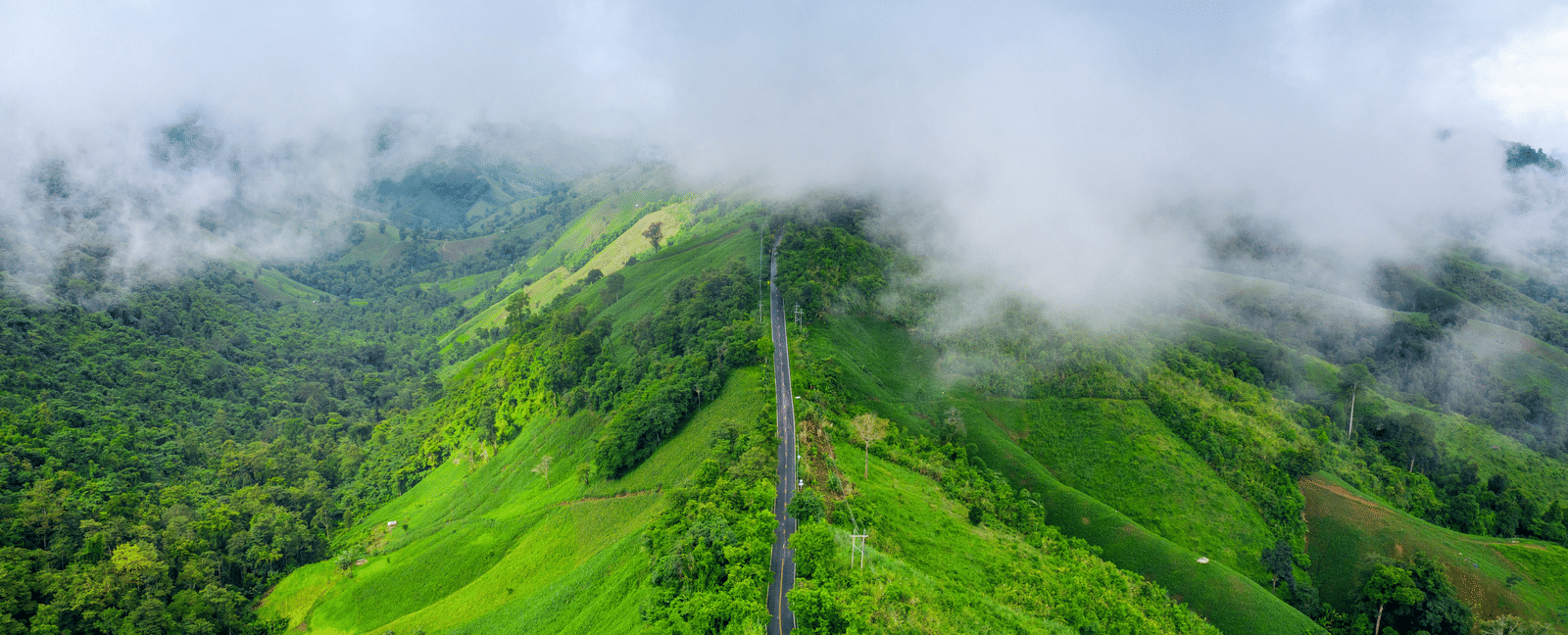 An aerial view of a winding mountain road cutting through lush hills, partially shrouded in low-hanging clouds