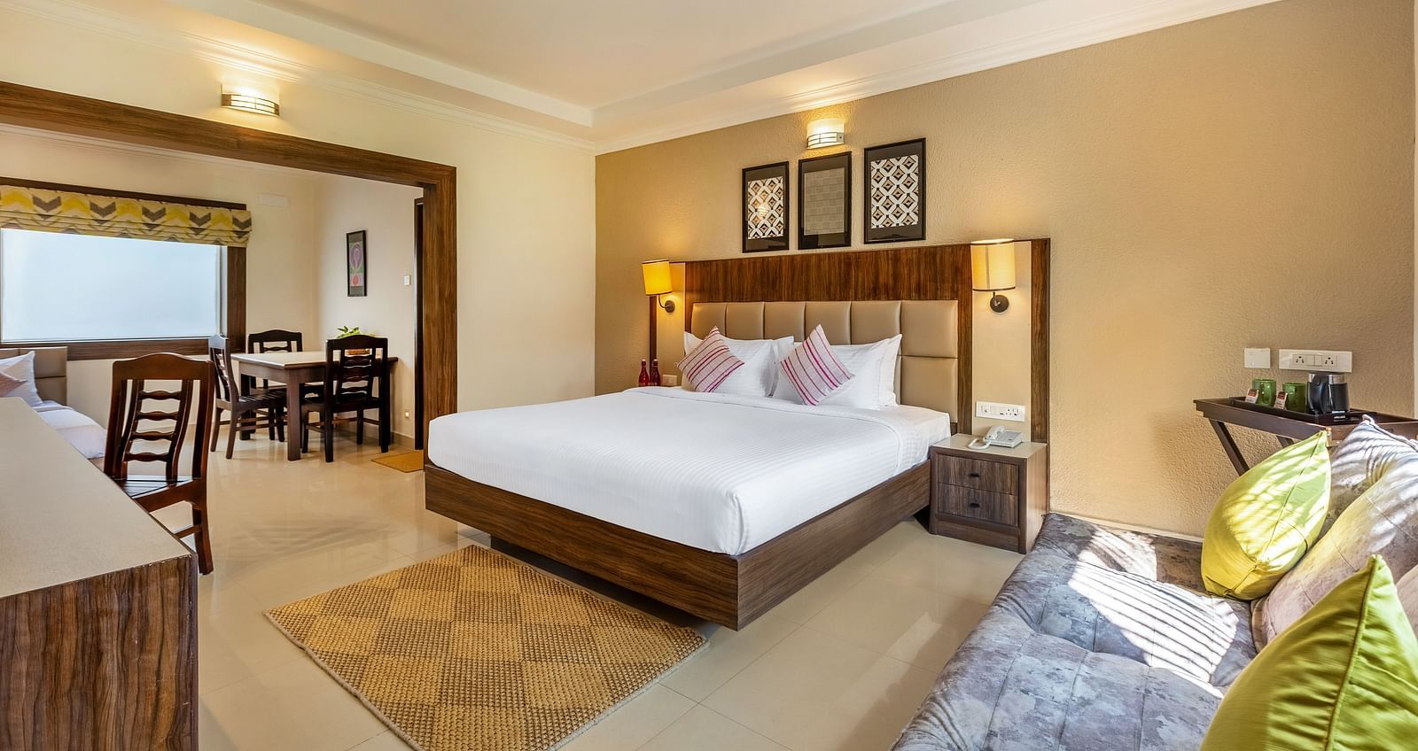 A hotel room with a queen bed, a sofa, and a doorway leading to a dining area with a table and chairs - Mango Hill Resort, Coimbatore