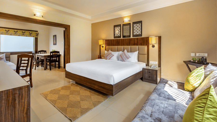 A hotel room with a queen bed, a sofa, and a doorway leading to a dining area with a table and chairs - Mango Hill Resort, Coimbatore