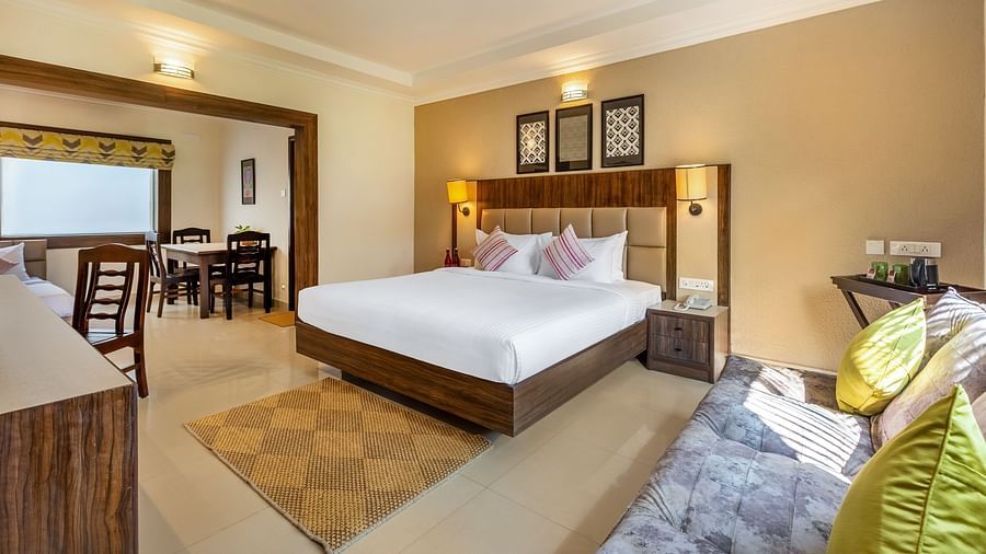 A hotel room with a queen bed, a sofa, and a doorway leading to a dining area with a table and chairs - Mango Hill Resort, Coimbatore