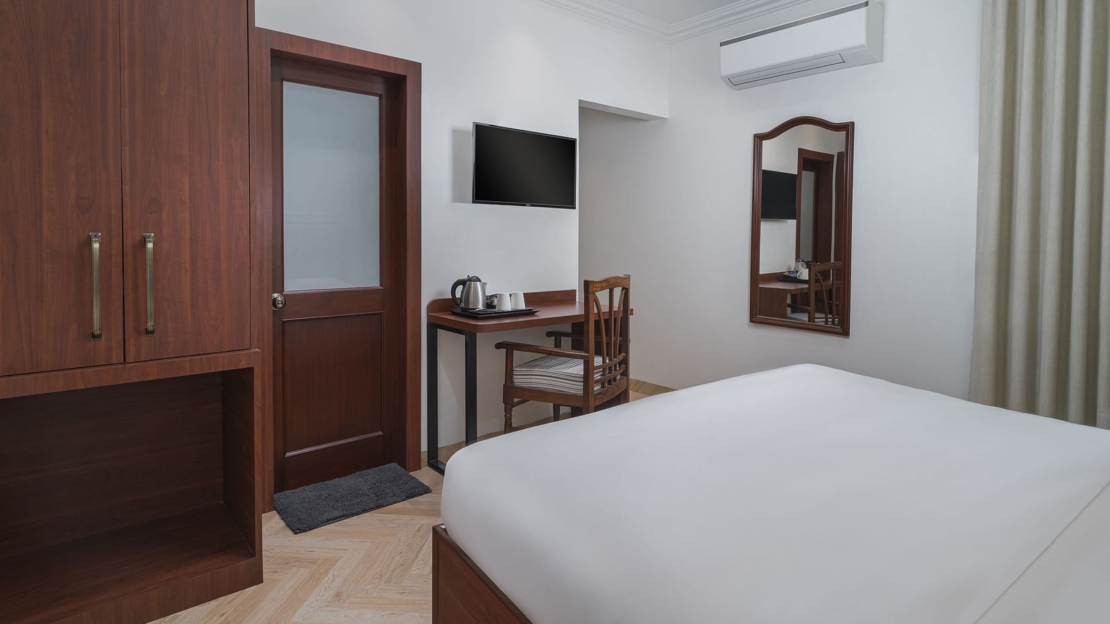 A room with a spacious bed, a wooden wardrobe, a wall-mounted television, and a wooden table directly below it | Hotel Ganga Maiya
