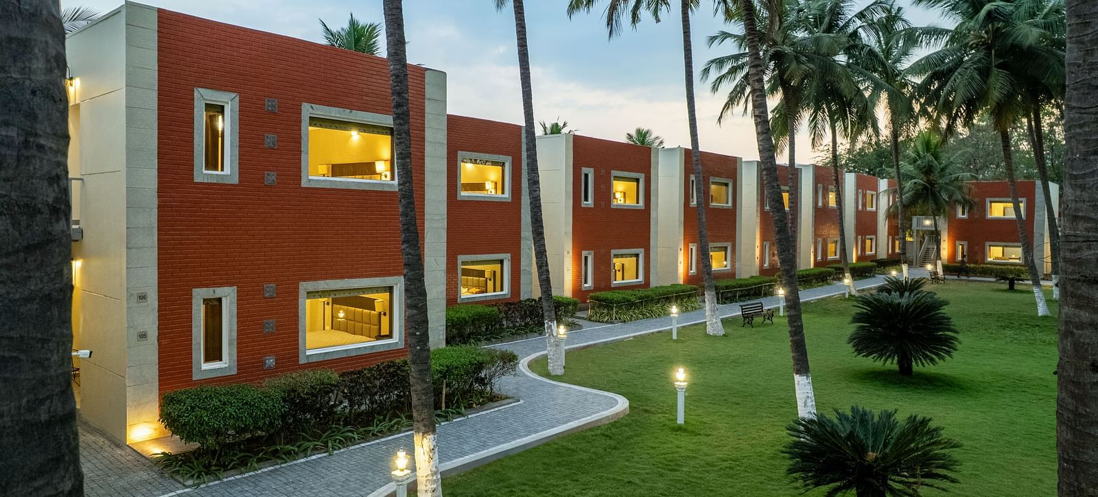 A building with red brick accents and white trim, surrounded by palm trees and a paved pathway - Mango Hill Resort, Coimbatore