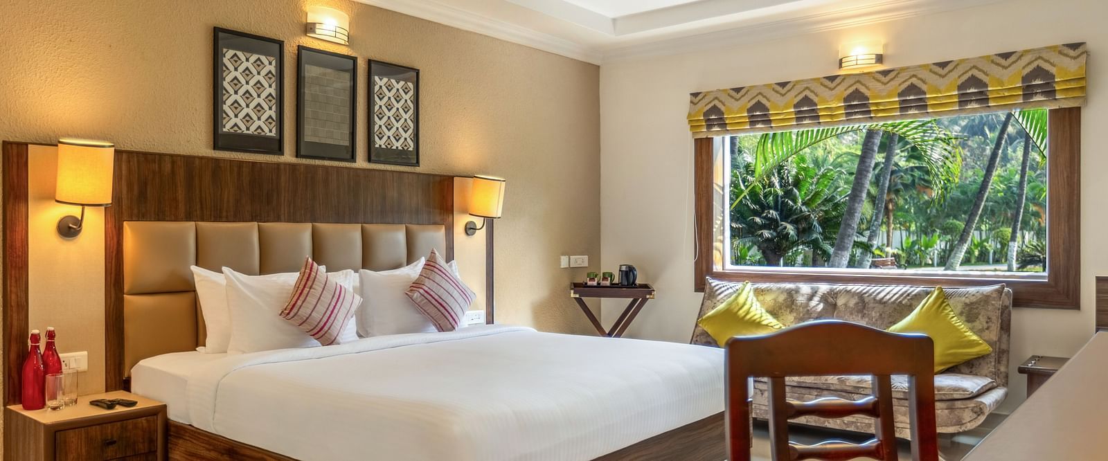 A hotel room with a bed, a desk, a sofa, and a large window overlooking a garden with palm tree - Mango Hill Resort, Coimbatore