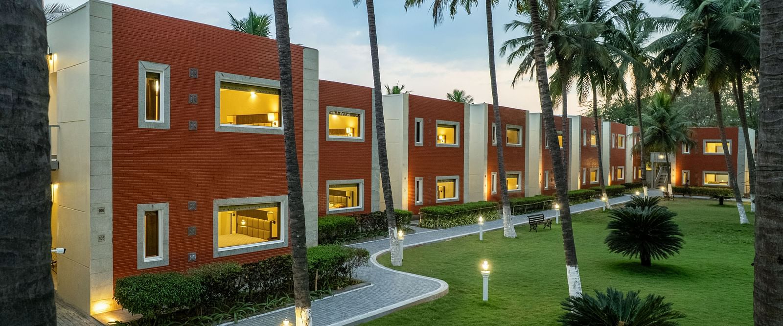 A building with red brick accents and white trim, surrounded by palm trees and a paved pathway - Mango Hill Resort, Coimbatore