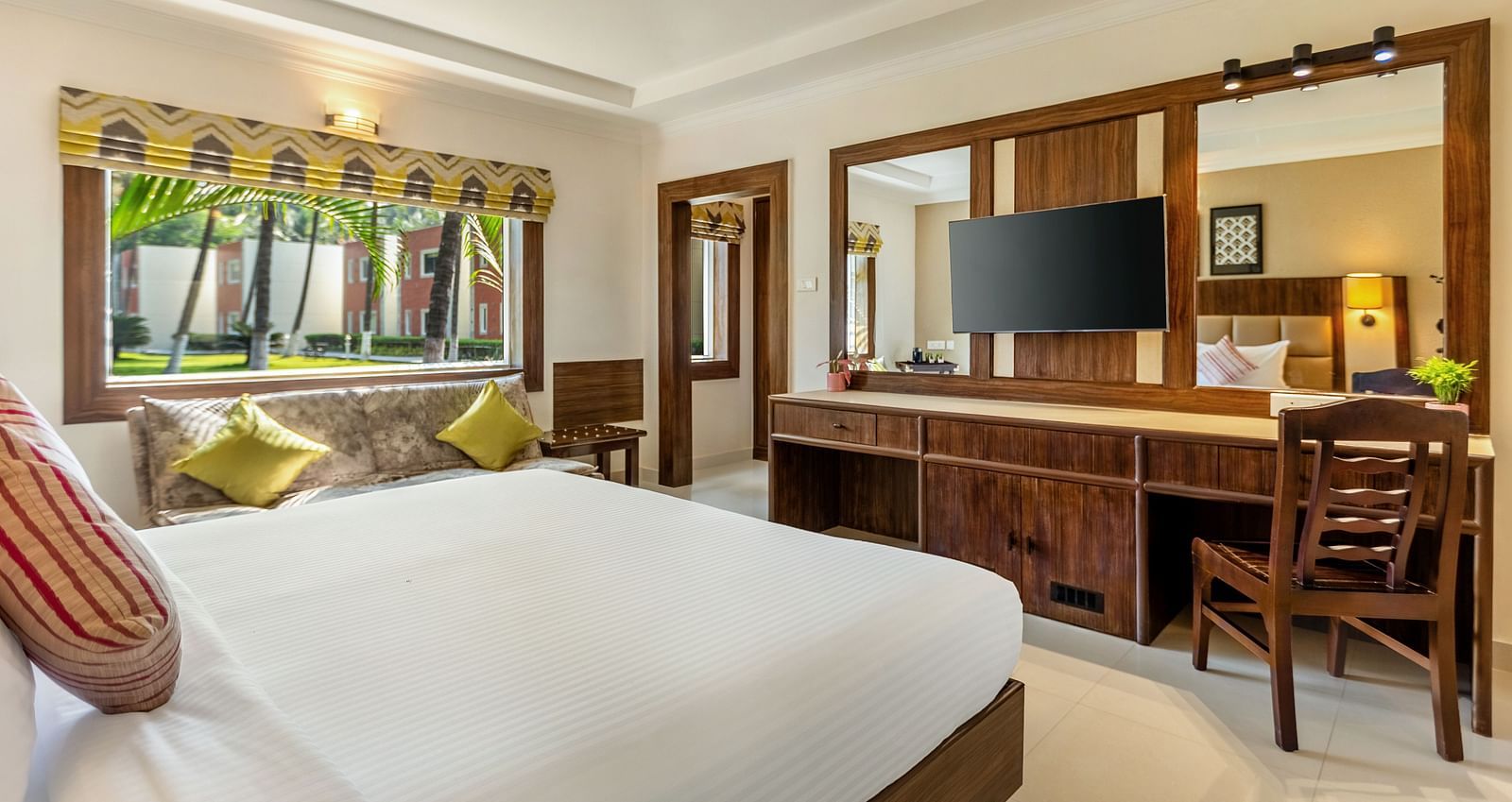 A bright bedroom with a bed with white linens, a wooden desk with a mounted TV, a patterned sofa with yellow pillows, and a window view of a tropical garden - Mango Hill Resort, Coimbatore