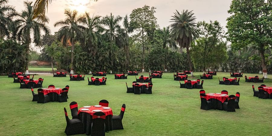 Outdoor lawn setup with round tables covered in red and black linen, surrounded by palm trees and greenery under a cloudy sky at DGV Resort by Treat, Silvassa