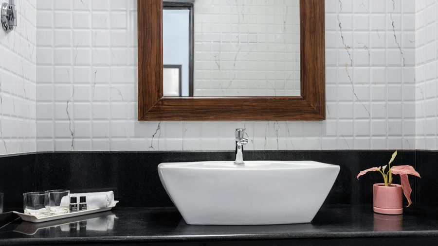 A bathroom with a white rectangular sink, a wooden framed mirror, white tiled walls, and a black countertop - Mango Hill Resort, Coimbatore