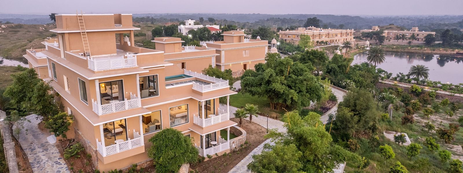 An image of our wellness retreat covered in lush green trees and a sky painted by the hue of the sun in the background - Rajbagh Wellness, Udaipur