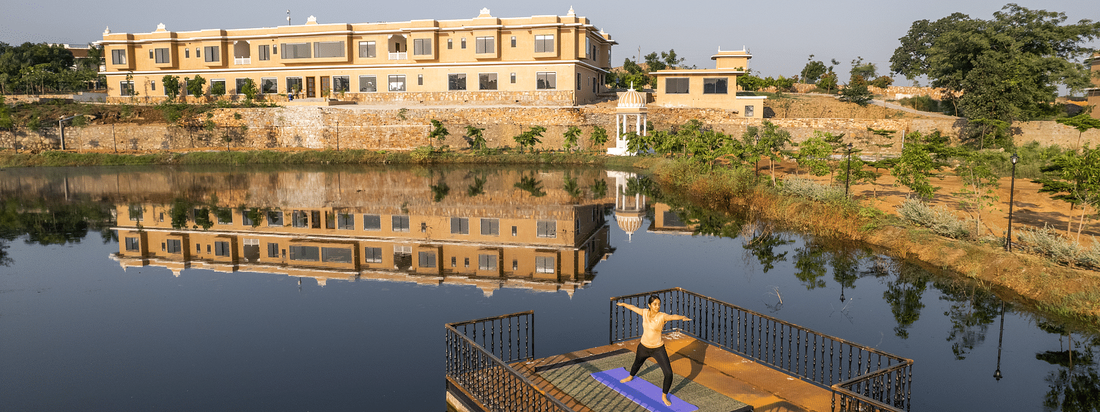 An image of a woman performing a yoga asan on a platform surrounded by water with our property facade in the back - Rajbagh Wellness, Udaipur
