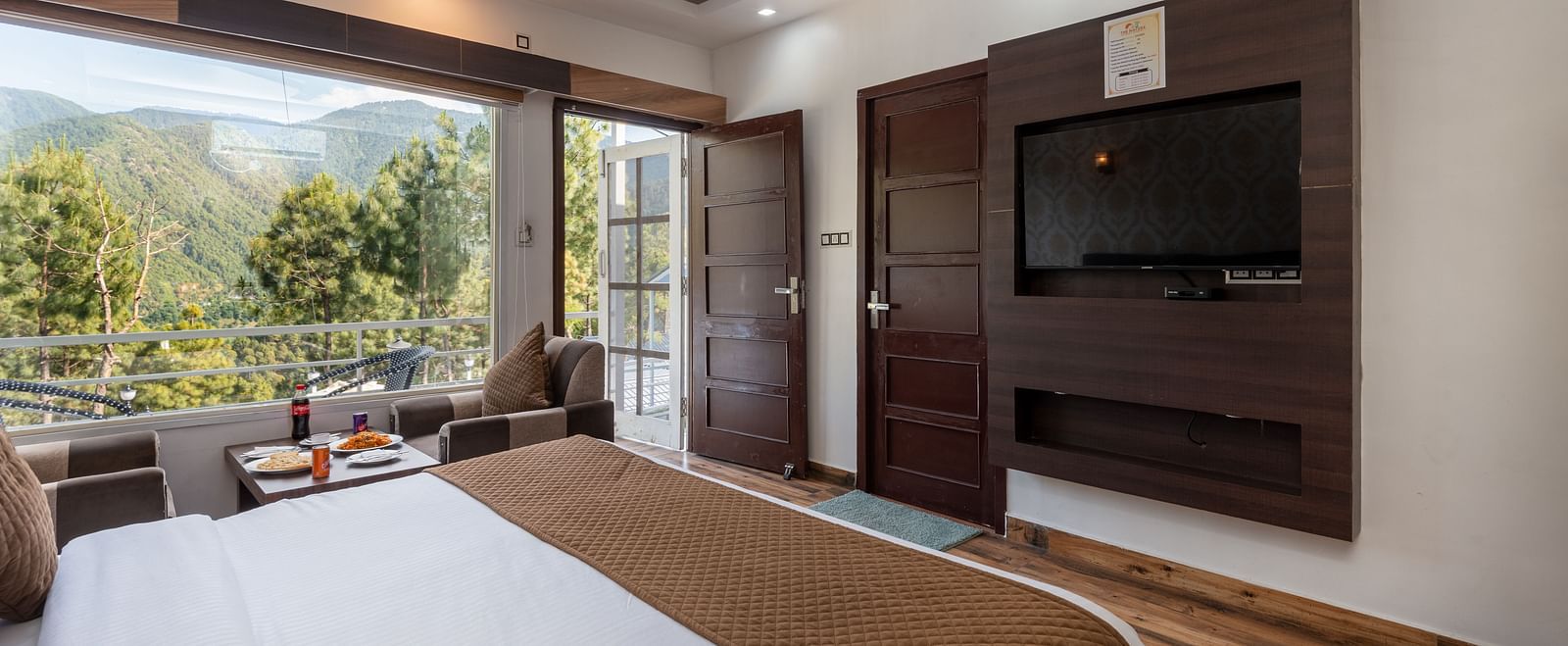  Cosy hotel room with a double bed, wall-mounted TV, wooden flooring, and an open door leading to a balcony with a scenic mountain and forest view - DLS Nature Trinket Resort, Dalhousie.