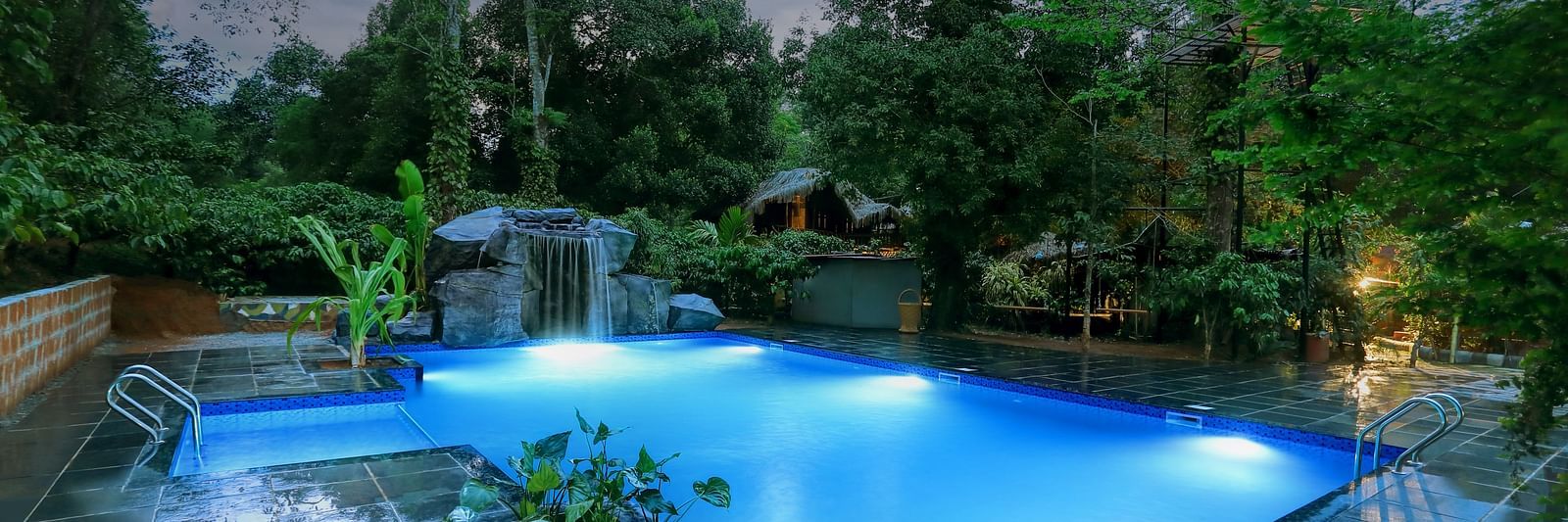 Illuminated resort pool at night with a waterfall and surrounding lush foliage - Evergreen County