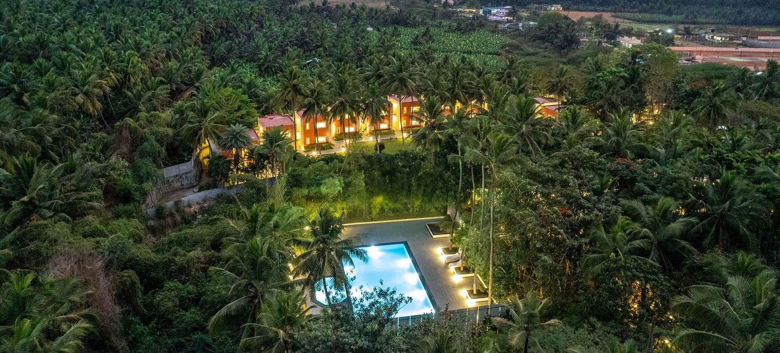 An aerial view of Mango Hill Resort, Coimbatore, nestled among palm trees, featuring a swimming pool and buildings with warm lighting, with mountains in the background.