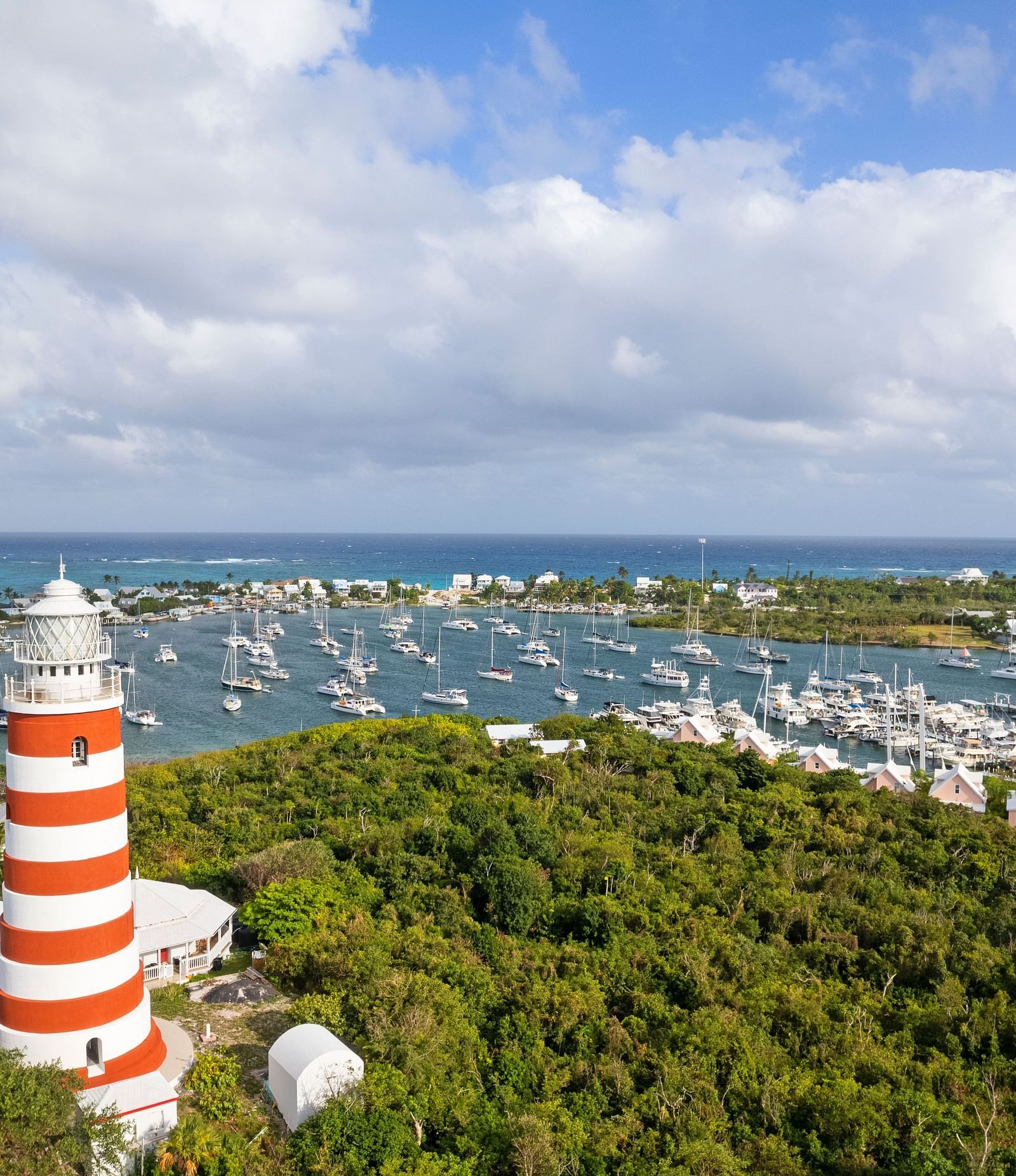 An aerial view of Hope Town, showing a red-and-white striped lighthouse, marina, and buildings surrounded by greenery and blue waters | Abaco Inn