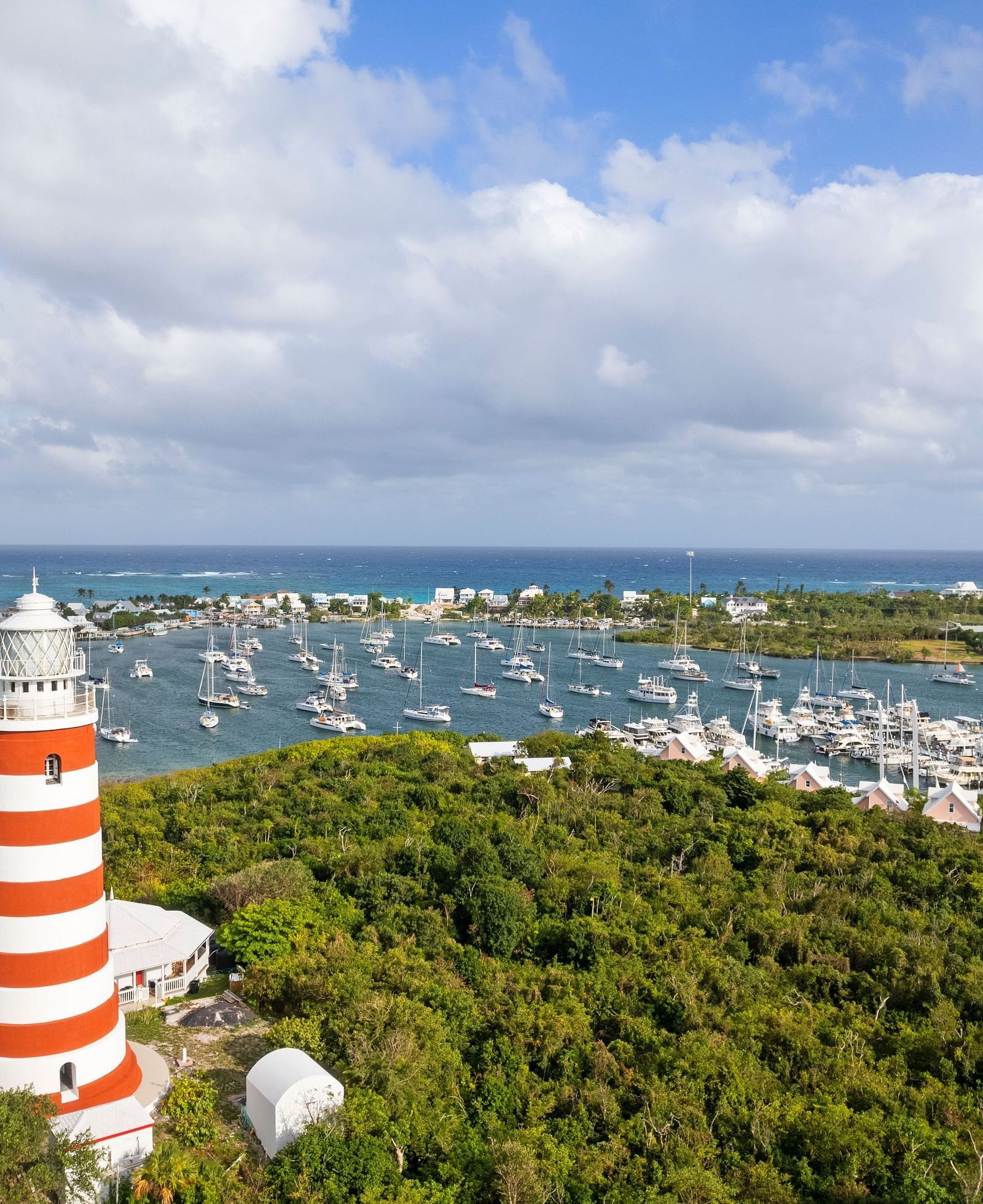 An aerial view of Hope Town, showing a red-and-white striped lighthouse, marina, and buildings surrounded by greenery and blue waters | Abaco Inn