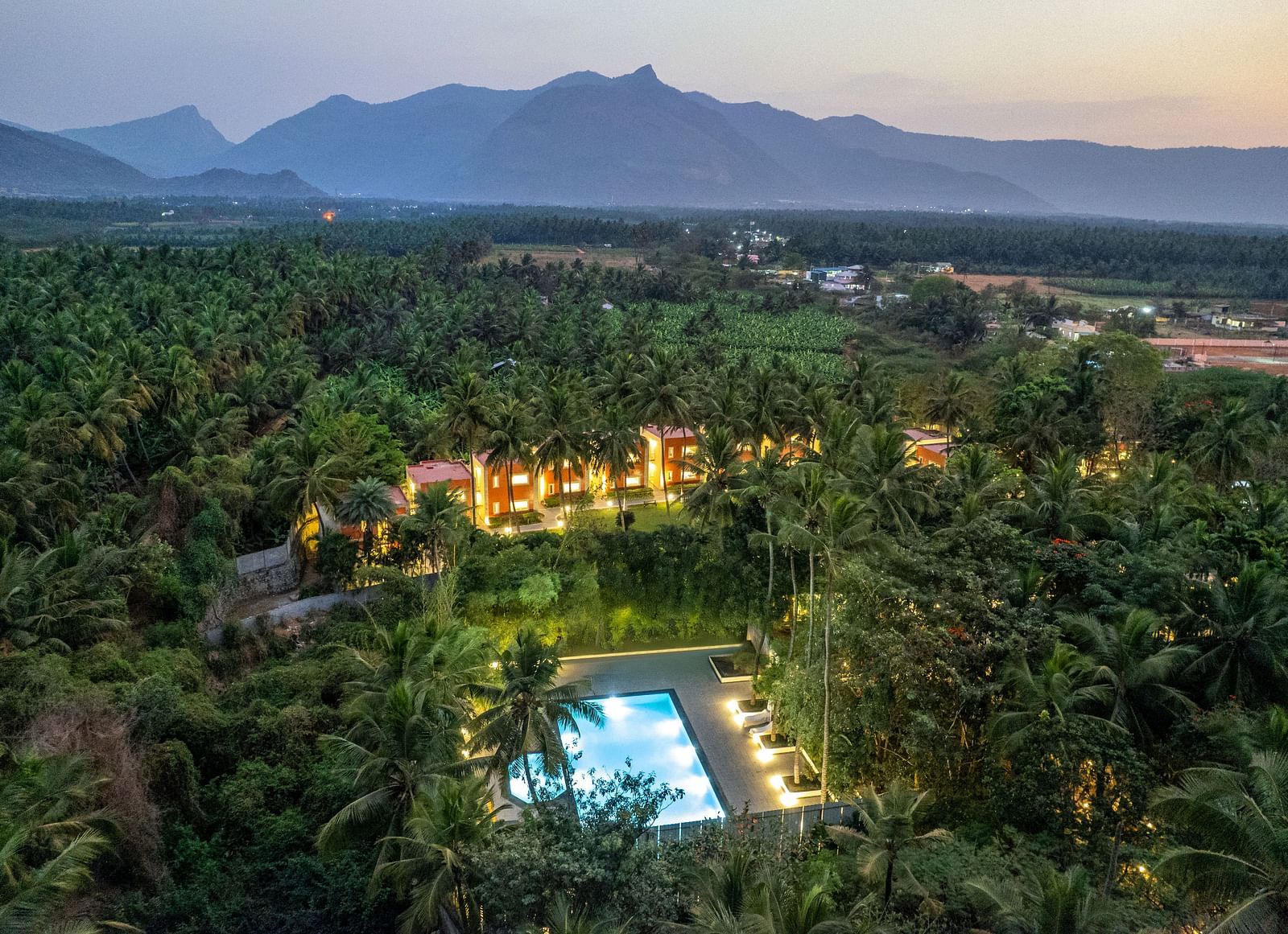 An aerial view of Mango Hill Resort, Coimbatore, nestled among palm trees, featuring a swimming pool and buildings with warm lighting, with mountains in the background.
