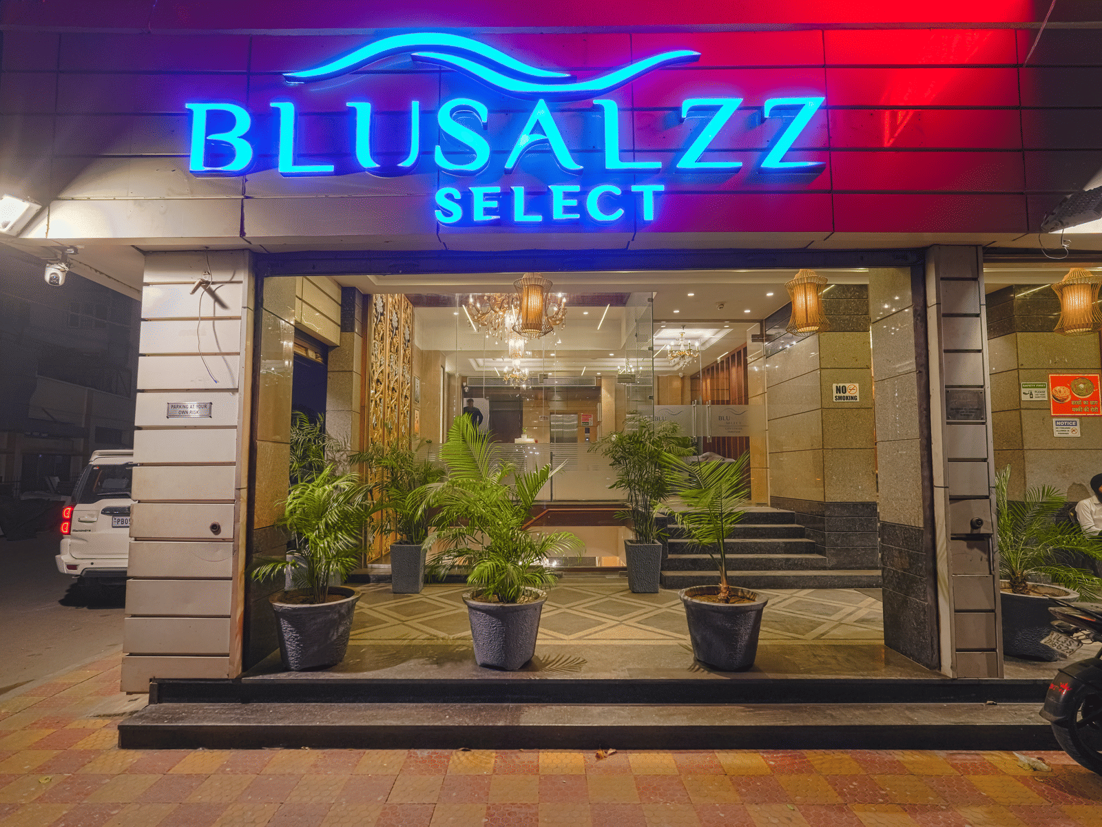 The ground-level entrance of the hotel at night, illuminated by a bright blue and red neon sign at BluSalzz Select City Centre, Amritsar.