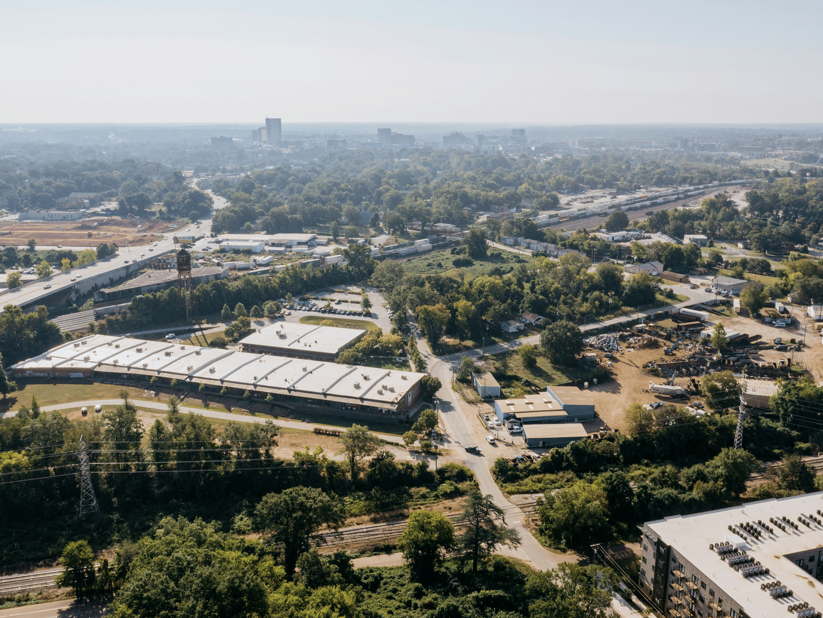 An aerial photo of an urban area with a large industrial building, a small city skyline in the distance, and extensive surrounding greenery and infrastructure like railroads and roads.