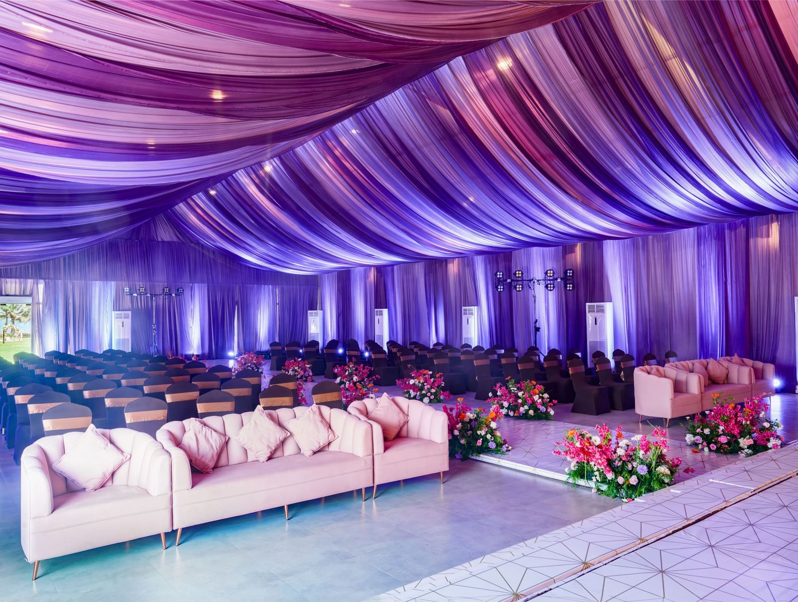 Decorated banquet hall with cream sofas and rows of chairs facing the stage featuring glowing purple decorated roof.