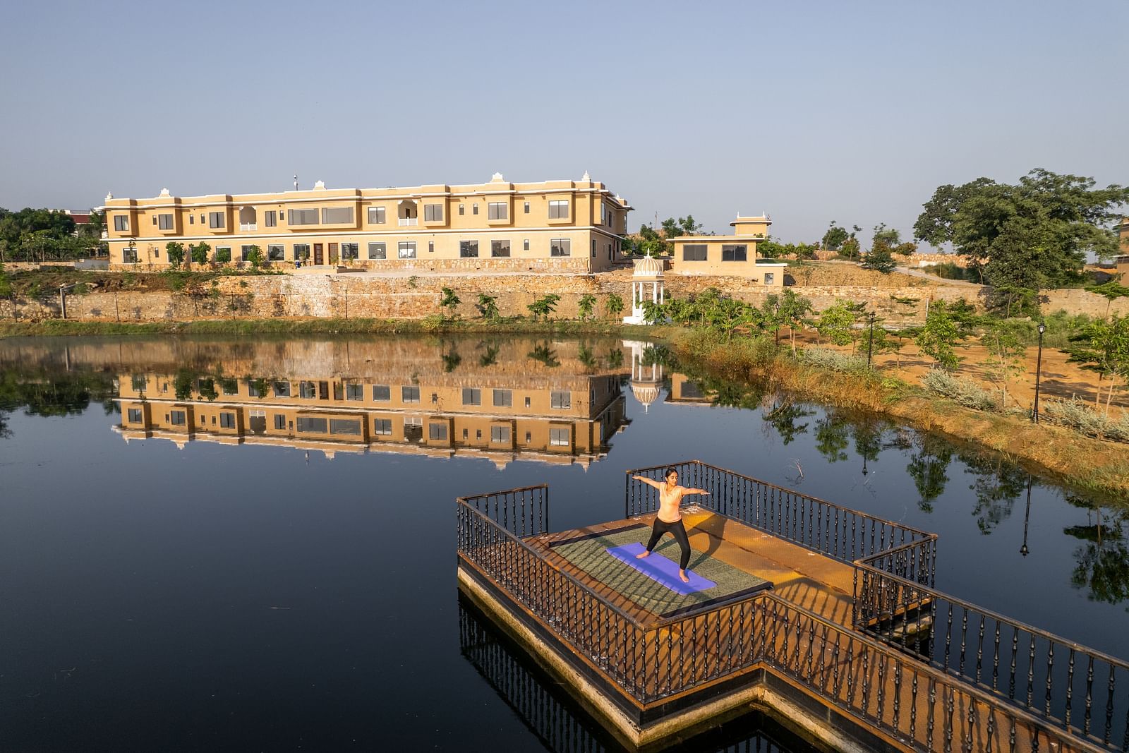 An image of a woman performing a yoga asan on a platform surrounded by water with our property facade in the back - Rajbagh Wellness, Udaipur