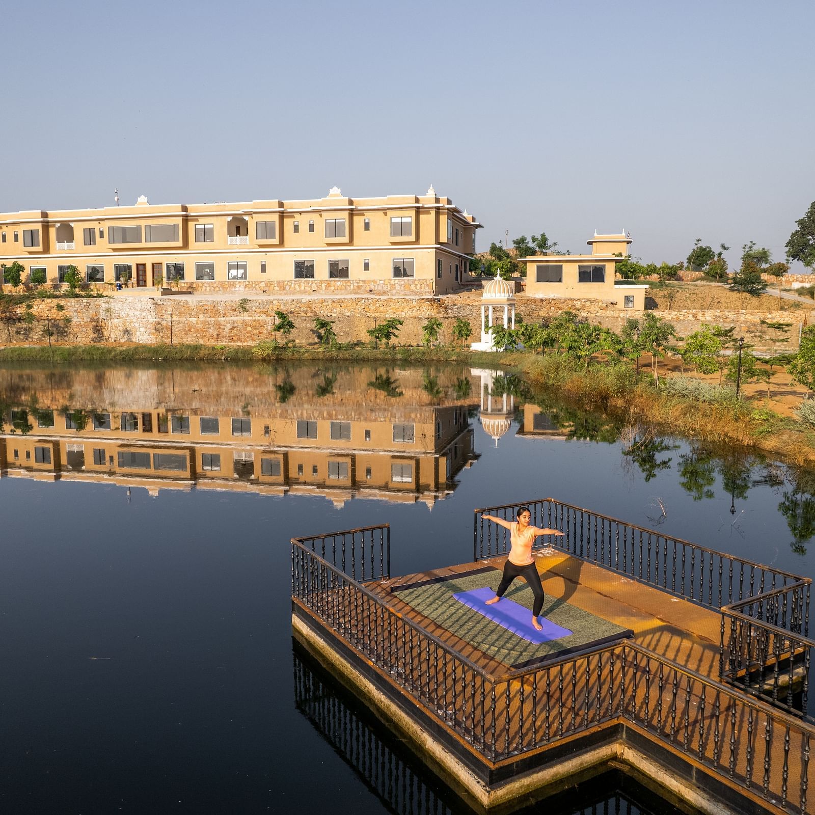 An image of a woman performing a yoga asan on a platform surrounded by water with our property facade in the back - Rajbagh Wellness, Udaipur