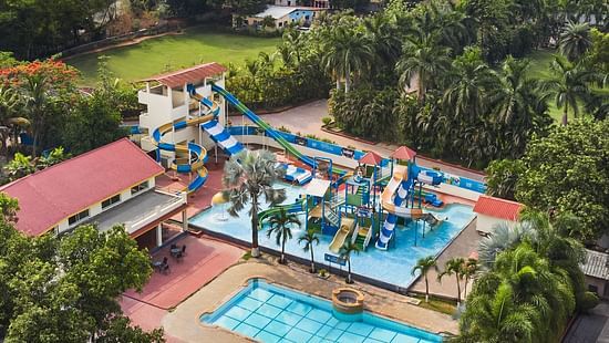 DGV Resort by Treat, Silvassa with a colourful waterpark featuring slides, splash areas, a swimming pool, and red-roofed buildings near a riverside