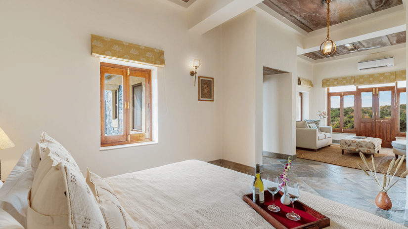 A king bed with a tray of that consists of a bottle of bubbly drink and champagne glasses on the bed inside one of the rooms - two chairs and a small table in between them on a balcony inside a room - Chunda Shikar Oudi, Udaipur