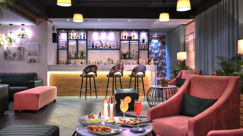 An image with a coffee table with food in the foreground and a bar counter with high chairs in the background  - Rooftop in Agra - Golden Tulip, Agra