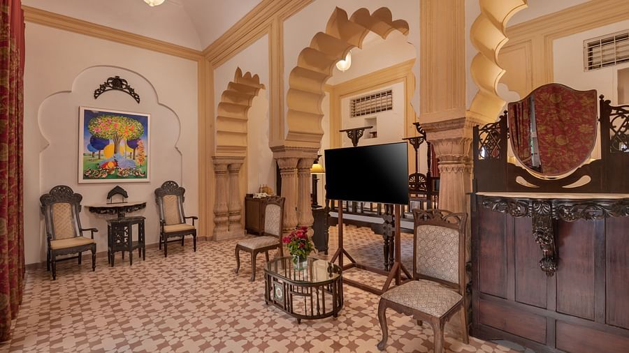 A room with chairs, tables, a television, a large cabinet, patterned flooring, and arched architectural details at Tijara Fort-Palace - 19th Century, Alwar.
